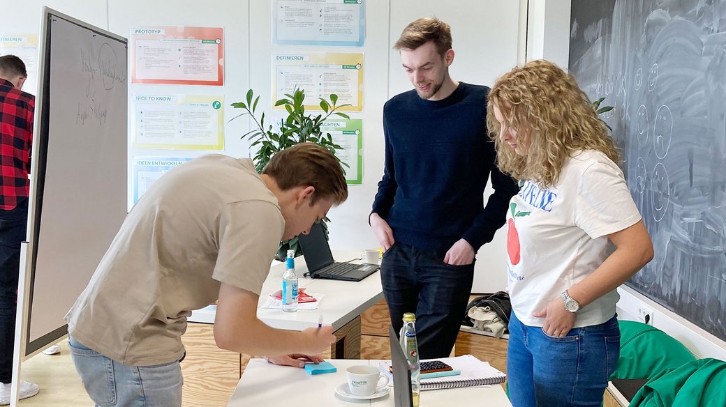 Drei Menschen schauen gemeinsam auf einen Tisch, wo einer der drei auf ein Post-it schreibt